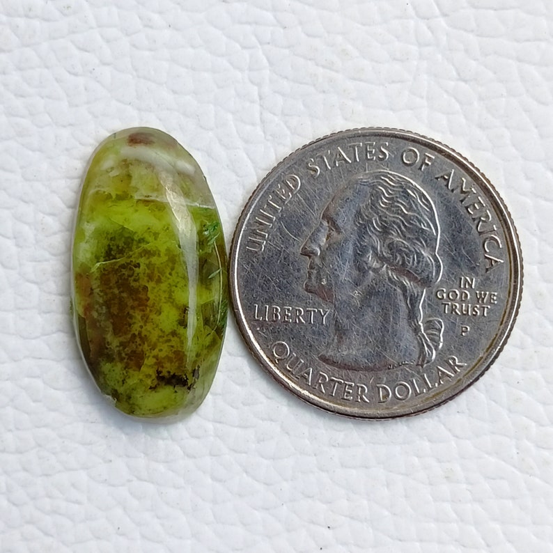 May include: An oval-shaped gemstone with a mix of green and brown hues. The stone is next to a quarter coin for size comparison. The gemstone has a polished, glossy surface and appears to be a natural stone.
