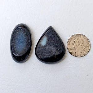 Op de afbeelding: Twee zwarte cabochon edelstenen met een witte, iriserende glans. De stenen zijn ovaal en traanvormig. Een Amerikaanse kwart dollar is opgenomen ter groottevergelijking.