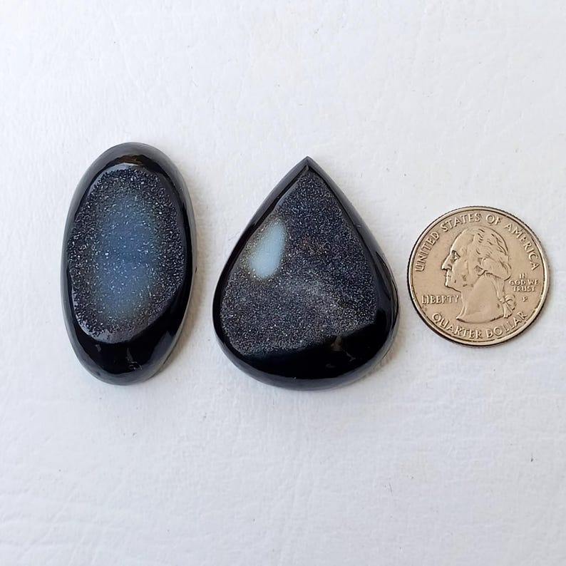 Op de afbeelding: Twee zwarte cabochon edelstenen met een witte, iriserende glans. De stenen zijn ovaal en traanvormig. Een Amerikaanse kwart dollar is opgenomen ter groottevergelijking.