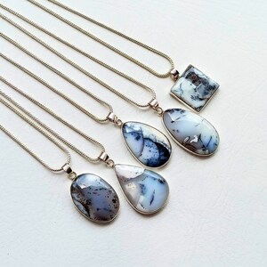 May include: A collection of silver necklaces with pendants. The pendants feature polished, natural stones in various shapes, including oval, teardrop, and square. The stones display a mix of white, gray, and black patterns. The necklaces are displayed on a white surface.