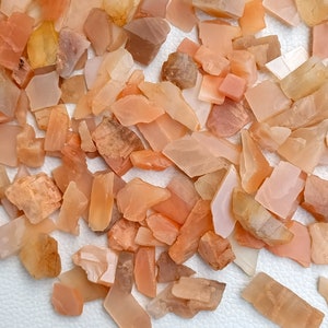 May include: A collection of small, irregularly shaped, orange sunstone chips. The chips are translucent and have a slight shimmer.