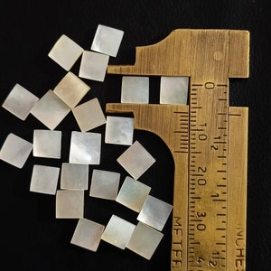 May include: A close-up of a ruler measuring a collection of white, iridescent, mother-of-pearl tiles. The tiles are square and have a slightly textured surface.