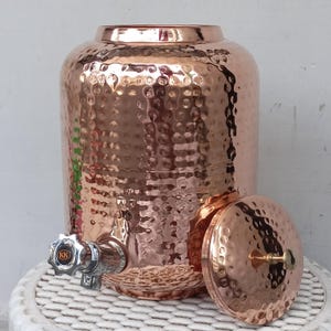 Pode incluir: Um dispensador de água de cobre com textura martelada, uma torneira prateada e uma tampa a condizer. O dispensador é cilíndrico com uma base larga e um topo ligeiramente mais estreito. A tampa está de lado, revelando o interior.