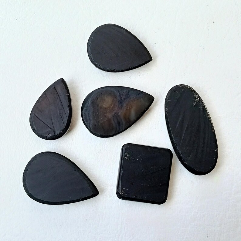 Op de afbeelding: Zes zwarte agaat cabochons in verschillende vormen, waaronder druppel- en vierkante vormen. De stenen hebben een gladde, gepolijste afwerking en een donkere, ondoorzichtige kleur.