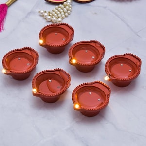 May include: Six terracotta-colored diya lamps with LED lights. The diyas have a decorative beaded edge and are arranged on a white surface.