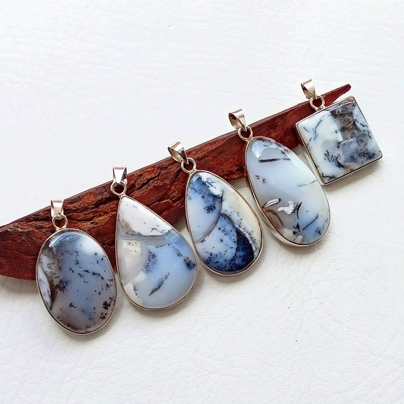 May include: Five dendritic opal pendants with silver-colored settings. The pendants vary in shape, including oval, teardrop, and square. Each features a unique pattern of white, gray, and black inclusions, creating a natural, artistic design. The pendants are displayed on a wooden surface.
