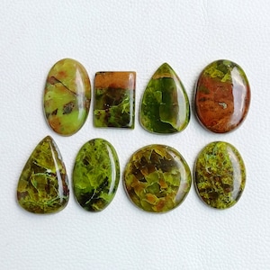 May include: Eight polished gemstones in various shapes, including oval, rectangular, and teardrop. The stones display a mix of green and brown hues, with a cracked, textured appearance. The gemstones are arranged on a white surface.