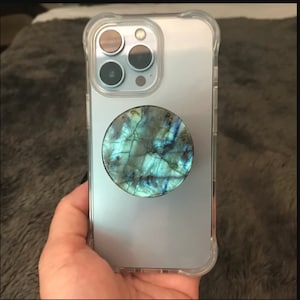 May include: A clear phone case with a silver phone inside. The phone has a black camera lens with three lenses. A round, blue and green, iridescent phone grip is attached to the back of the phone.
