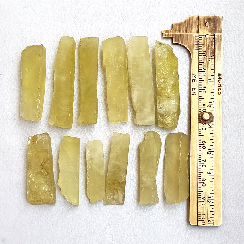 Puede incluir: Doce piezas de cristal de calcita amarillo verdoso, cada una de aproximadamente 2,5 cm de largo, dispuestas en dos filas sobre una superficie blanca. Se incluye una regla para la escala.