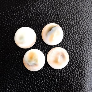 Op de afbeelding: Vier ronde, witte, iriserende schelpcabochons met bruine en grijze markeringen. De cabochons zijn in een vierkant op een zwart, getextureerd oppervlak gerangschikt.