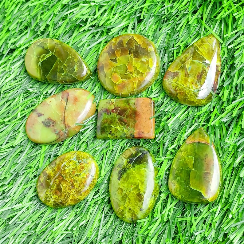 May include: A collection of eight polished gemstones in shades of green and brown, with various shapes including oval, teardrop, and rectangular. The gemstones are displayed on a bed of green artificial grass.