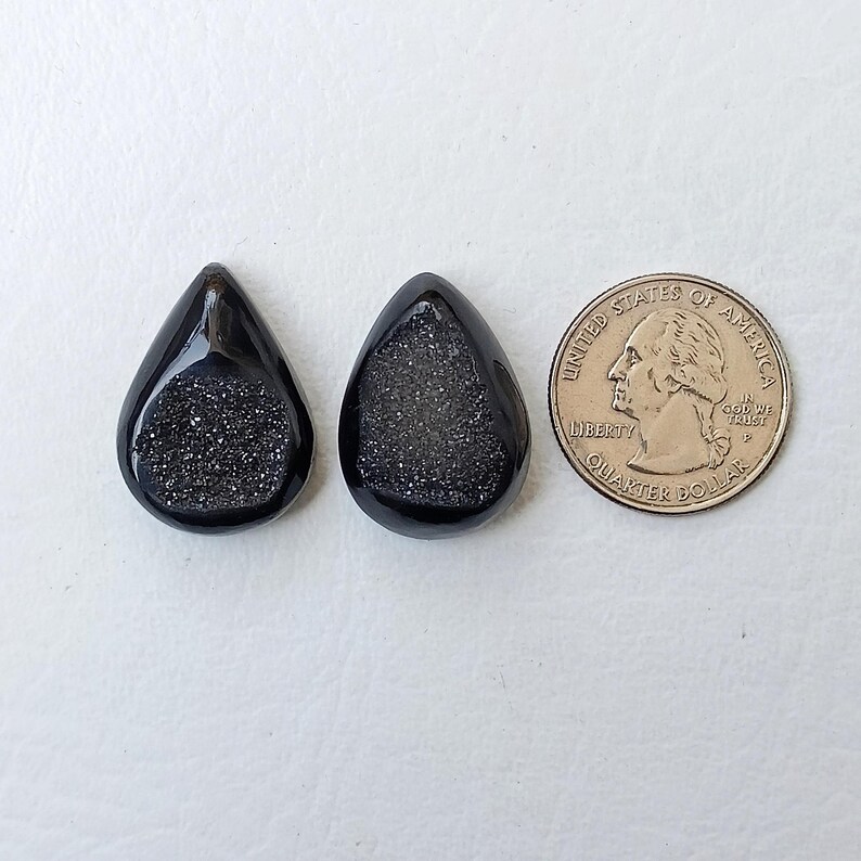 Op de afbeelding: Twee zwarte druppelvormige edelsteencabochons met een glinsterend, getextureerd oppervlak.