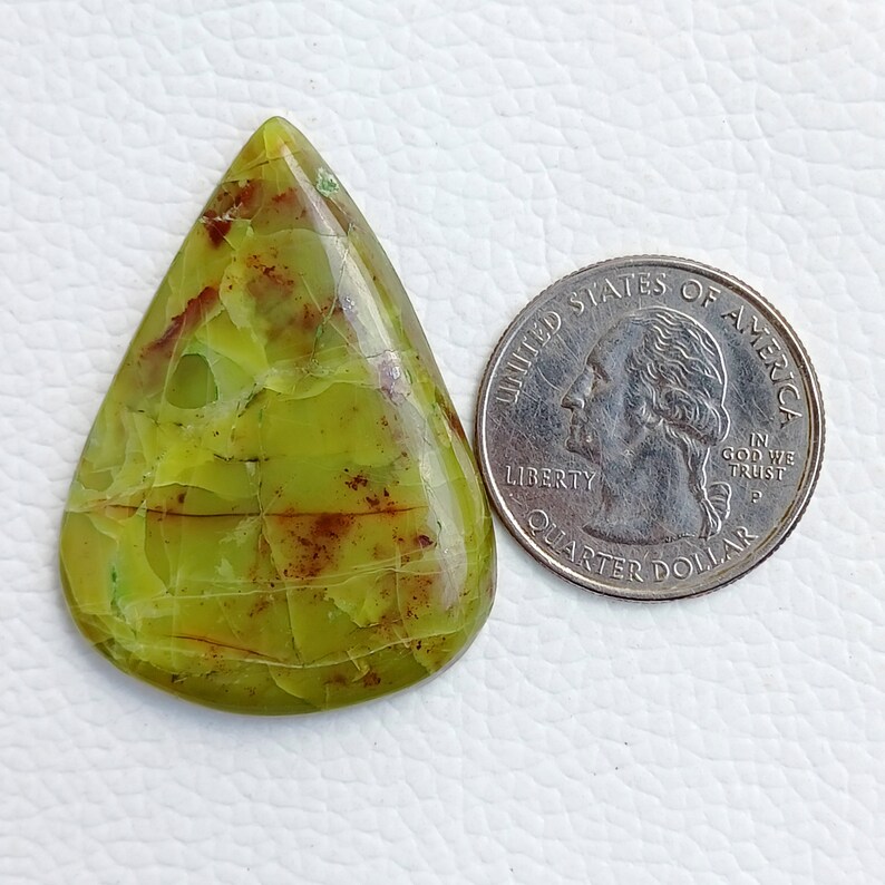 May include: A pear-shaped, polished gemstone with a light green hue and brown veining. A U.S. quarter coin is placed next to the gemstone for size comparison. The gemstone has a cracked appearance.