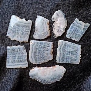 Puede incluir: Una colección de ocho rebanadas de ágata, cada una con un patrón único de bandas blancas y grises. Las rebanadas son lisas y pulidas, con una apariencia translúcida.