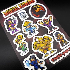 May include: A sheet of Mortal Kombat stickers featuring pixelated character designs. The stickers include characters in various poses, with the Mortal Kombat logo and the phrase "FINISH HIM!!" in red and white.