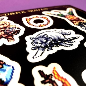 May include: A set of eight Dark Souls themed stickers featuring pixelated characters and objects from the video game. The stickers are printed on white paper and have a black background.