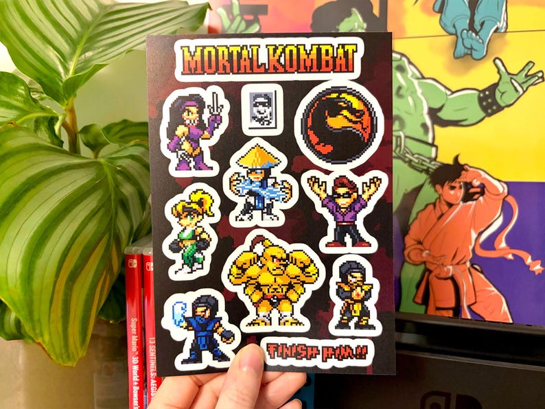 May include: A sheet of pixel art stickers featuring characters and the logo from the video game Mortal Kombat. The stickers include characters like Scorpion, Sub-Zero, and Sonya Blade. The words "Mortal Kombat" and "Finish Him!!" are visible.