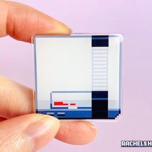 May include: A clear acrylic pin featuring a pixelated design of a video game console. The console is white with a blue and black control panel and a red power light.