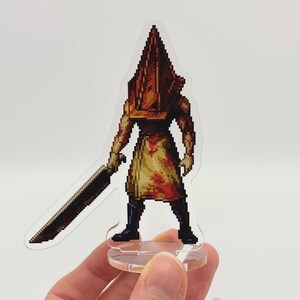 May include: A pixelated acrylic figure of a character with a pyramid-shaped head, holding a large sword. The figure is in shades of brown, red, and yellow, with a clear base. The figure is a video game character.