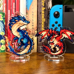 May include: Two pixel art dragon figurines, one blue and one red, on clear circular stands. The background features books and a blue games console controller. The figurines are made of acrylic.