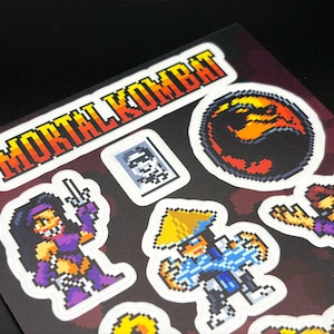 May include: Sheet of pixel art stickers featuring characters and logos from the video game Mortal Kombat. The stickers include the game's logo, a dragon symbol, and various character illustrations in a retro style.