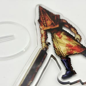 May include: A clear acrylic stand featuring a pixelated image of a character with a pyramid-shaped head, holding a large blade. The character is depicted in shades of brown, yellow, and red. A separate, round clear base is included.