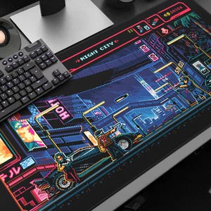 May include: A black rectangular gaming mouse mat with a pixelated cityscape design. The design features neon colours, a futuristic aesthetic, and the words "NIGHT CITY". A black keyboard and speaker are also visible.