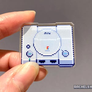 May include: A white and grey pixel art sticker of a PlayStation console. The sticker is clear and has a glossy finish.