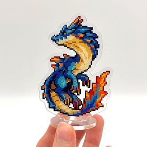 May include: A pixelated dragon figurine in shades of blue, orange, and yellow. The dragon has a flame-like tail and is mounted on a clear stand. The design is reminiscent of retro video game graphics.