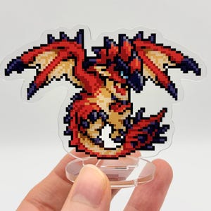 May include: A pixelated dragon figurine in red, orange, and blue hues. The dragon has outstretched wings and is mounted on a clear, circular base. The figurine is held in a person's hand.