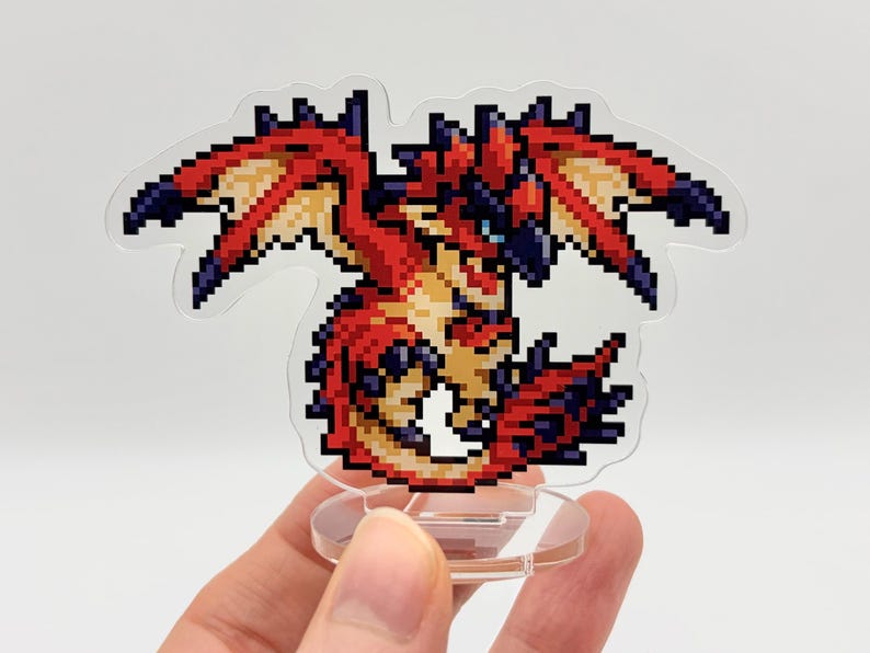 May include: A pixelated dragon figurine in red, orange, and blue hues. The dragon has outstretched wings and is mounted on a clear, circular base. The figurine is held in a person's hand.