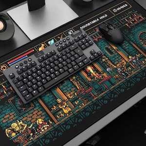 May include: A black gaming mouse and keyboard sit on a large, rectangular mousepad. The mousepad features a colorful pixel art design of a dungeon scene with the text "ROUNDTABLE HOLD" and "TARNISHED". A black speaker is visible in the background.