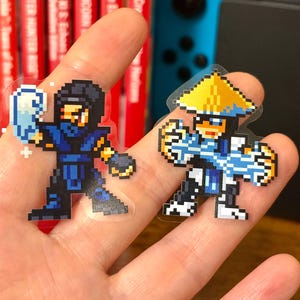 May include: Two pixel art stickers featuring characters from a fighting game. One character is in a blue and black outfit with a mask and holding an ice blast. The other character is in a white and blue outfit with a yellow hat.