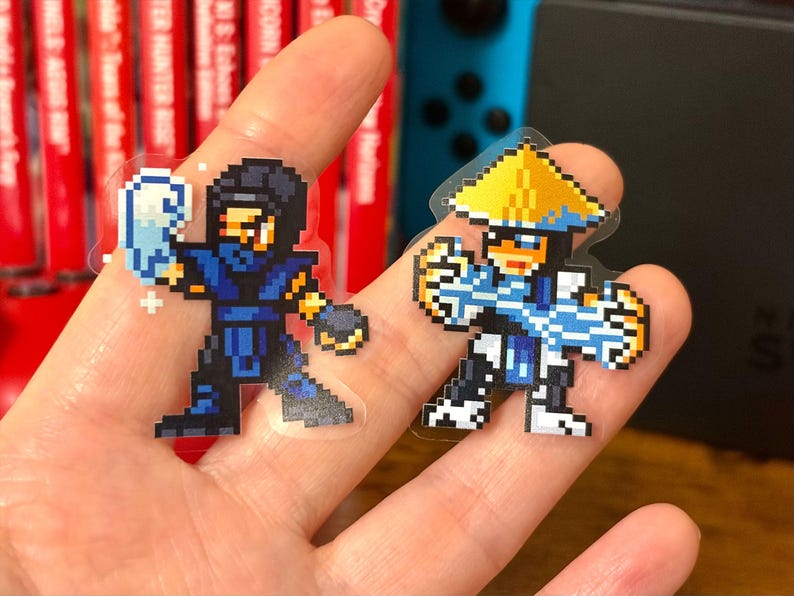 May include: Two pixel art stickers featuring characters from a fighting game. One character is in a blue and black outfit with a mask and holding an ice blast. The other character is in a white and blue outfit with a yellow hat.
