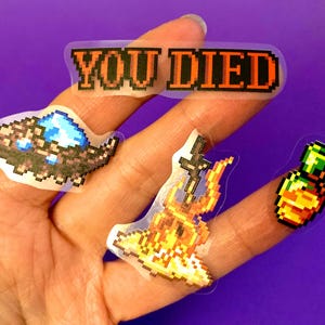 May include: Four pixel art stickers on a hand. The stickers are on a clear background. The stickers are of a sword and fire, a bottle of potion, a creature with a blue gem, and a banner that says "YOU DIED".
