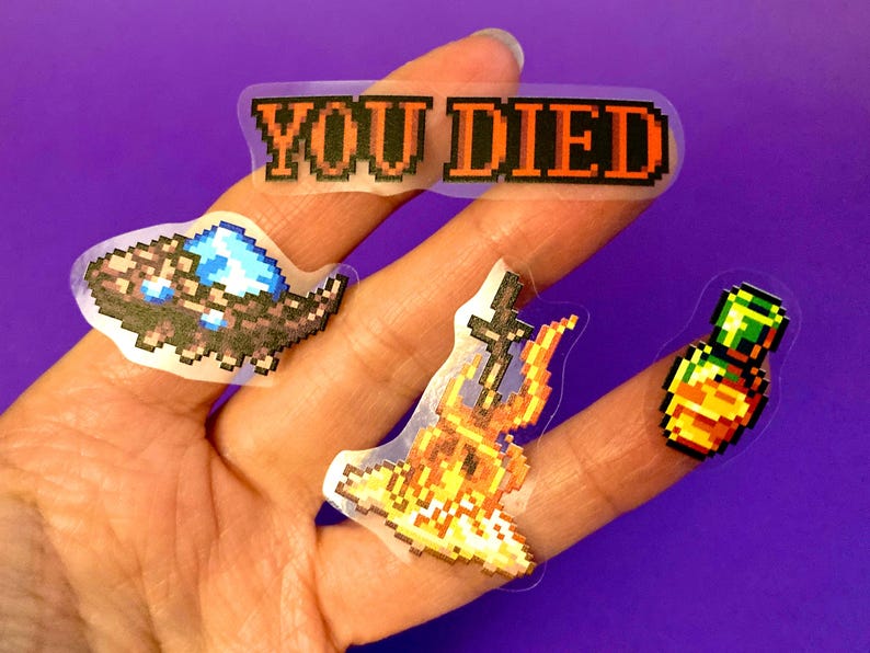 May include: Four pixel art stickers on a hand. The stickers are on a clear background. The stickers are of a sword and fire, a bottle of potion, a creature with a blue gem, and a banner that says "YOU DIED".