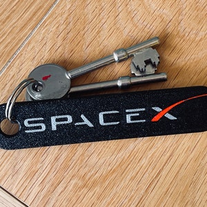 Puede incluir: Un llavero negro de SpaceX con el logotipo de SpaceX en blanco y naranja, y dos llaves plateadas sobre una superficie de madera. El llavero tiene forma ovalada y una superficie texturizada.