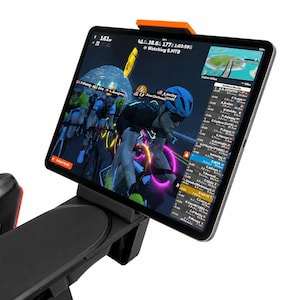 May include: A black tablet is mounted on a black and orange holder, displaying a cycling simulation with cyclists. The tablet is angled towards the viewer, showing a detailed interface with performance metrics and a virtual race environment.
