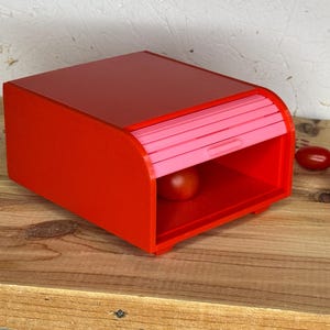May include: A vibrant red bread bin with a pink, roll-top door. A single tomato sits inside the box, with several more tomatoes scattered on the wooden surface beside it. The box has a rectangular shape with rounded edges.