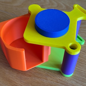 May include: A colorful, geometric toy with an orange container, a yellow platform, and a blue disc. The toy also features green, purple, and yellow cylindrical supports. The design is modern and playful.