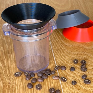 Könnte beinhalten: Ein transparenter Kaffeemühle mit einem schwarzen trichterförmigen Aufsatz. Zwei zusätzliche Trichter in Grau und Rot sind in der Nähe. Kaffeebohnen liegen auf der Holzoberfläche verstreut.
