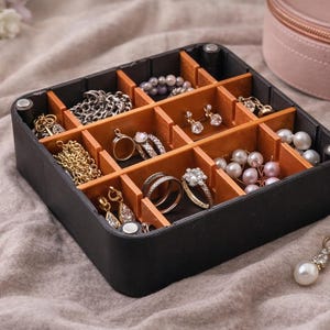 Peut inclure: Un organisateur de bijoux noir avec plusieurs compartiments remplis de divers bijoux. Les bijoux comprennent des colliers, des bagues, des boucles d'oreilles et des bracelets dans des tons or et argent, avec des perles et des pierres précieuses. Un étui à bijoux rond rose est en arrière-plan.