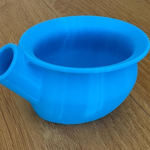 May include: A bright blue, round, plastic bowl with a spout on the side. The bowl has a wide rim and a smooth, curved surface. The spout is cylindrical and angled downward. The bowl is sitting on a wooden surface.