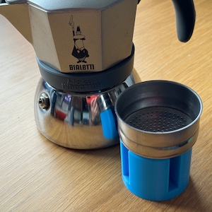 May include: A stainless steel Bialetti Moka Induction coffee maker with a black handle and blue accents. The filter basket is separate and has a blue base. The Bialetti logo is visible.