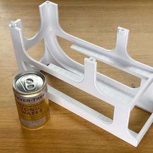 Pode incluir: Um organizador de plástico branco para latas de bebidas com dois níveis para armazenamento horizontal, posicionado ao lado de uma lata dourada e branca de Fever-Tree Premium Indian Tonic Water para mostrar a escala da unidade.