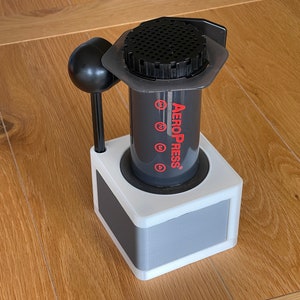 Peut inclure: Un appareil à café AeroPress noir et gris, posé dans un support blanc et gris. L'AeroPress porte des marques rouges et le nom de la marque en rouge. Un agitateur noir est positionné sur le côté. L'ensemble est sur une surface en bois.
