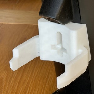 May include: A white 3D-printed bracket with a curved design, featuring a slot and a cross-shaped detail. The bracket is attached to a black surface and a dark brown object.