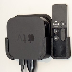 Può includere: Un supporto a parete nero per Apple TV e telecomando. L'Apple TV è nera con il logo Apple e la scritta "tv". Il telecomando è anch'esso nero con diversi pulsanti. Il supporto è progettato per mantenere i dispositivi organizzati.