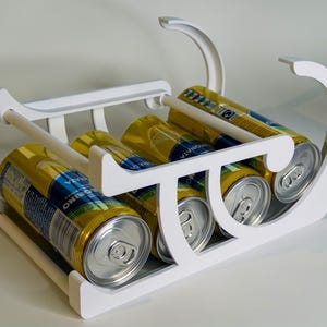 May include: A white, modern can holder filled with six gold and blue beverage cans. The holder has a rectangular base and curved handles, designed to carry and display canned drinks. The cans are arranged neatly within the holder.