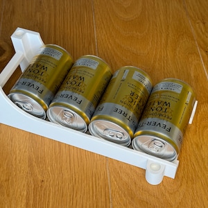 Può includere: Un portabevande in plastica bianca con quattro lattine dorate e argentate di Fever-Tree Indian Tonic Water. Le lattine sono disposte in fila, su una superficie di legno.